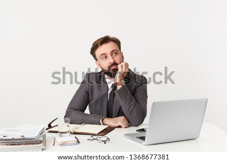 Bearded manager sits at the table props his chin with his hand looks to the right side bored doesn't know what to do thinks about something unimportant, dressed in a expensive suit with a tie