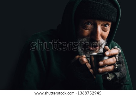 Old homeless man with grey beard covering up in green decrepit wear holding a mug of hot tea to warm himself in a cold night Royalty-Free Stock Photo #1368752945