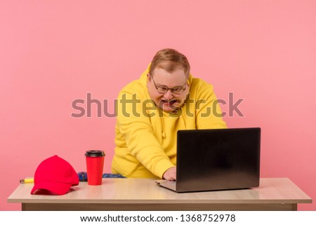 Education, work, overwork concept. Ridiculous, tired from work, mad man in yellow sports cloth look at monitor of a pc with comical foolish grimace, isolated on light pink background