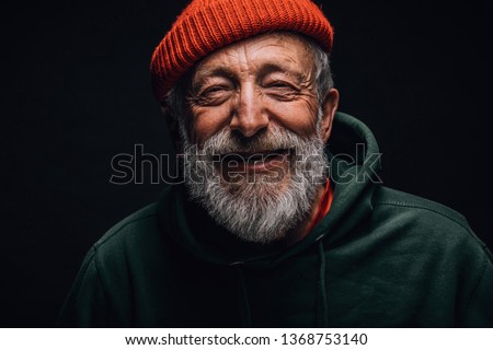 Close up portrait of happy 70-year-old optimist man with smiling wrinkled face, dressed in hipster orange hat and green hoodie, isolated over black background. Positive and cheerful at any age. Royalty-Free Stock Photo #1368753140