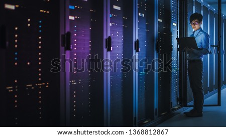 In Dark Data Center: Male IT Specialist Stands Beside the Row of Operational Server Racks, Uses Laptop for Maintenance. Concept for Cloud Computing, Artificial Intelligence, Supercomputer. Neon Lights Royalty-Free Stock Photo #1368812867