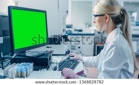 Female Research Scientist Sits at His Workplace in Laboratory, Uses Green Mock-up Screen Personal Computer. In the Background Genetics, Pharmaceutical Research Centre.