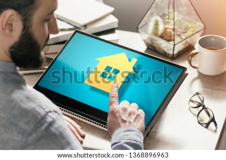 Bearded man with laptop at his desk. He presses on the house icon. Concept of real estate buying, booking, advertising via internet. Image