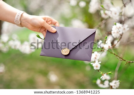 Close-up photo of female hands holding purple invitation envelope with wax seal, gift certificate,wedding invitation card on the background of blooming flowers
