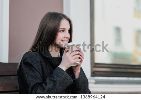 Young beautiful millenial girl in a coat is sitting on an outdor bench with a paper cup of coffee. Autumn or spring day, cool weather. The girl smiles, drinks the drink and takes pictures on the