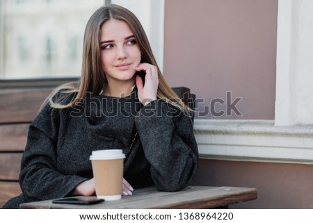 Young beautiful millenial girl in a coat is sitting on an outdor bench with a paper cup of coffee. Autumn or spring day, cool weather. The girl smiles, drinks the drink and takes pictures on the