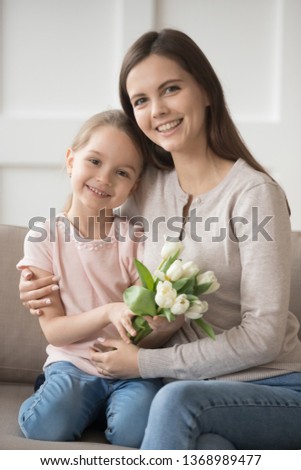Vertical image family portrait mum hugs small daughter holds white tulips flowers sitting on sofa at home, kid make present for loving mom. Mother day birthday life events holidays celebration concept