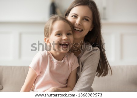 Happy preschool daughter kid sitting on mother lap on sofa in living room older younger sisters laughing looking at camera feels overjoyed positive emotions, posing for picture family portrait concept