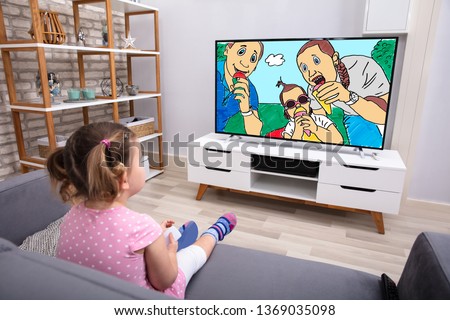 Rear View Of Innocent Girl Sitting On Sofa Watching Cartoon On Television