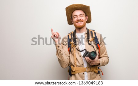 Young redhead explorer man pointing to the side with finger. Holding a camera.