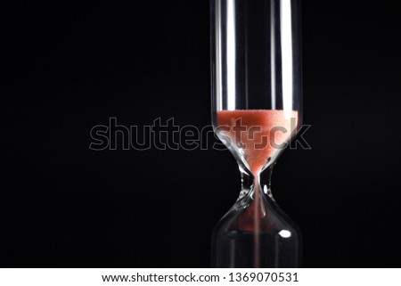 Macro picture of small modern hourglass against solid black background with coral sand trickling. Hard light spot on the right.