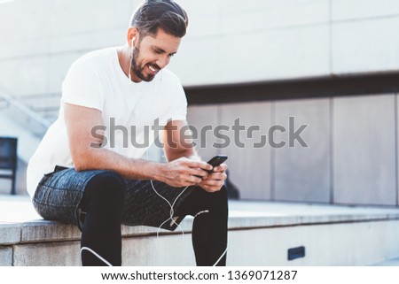 Happy smiling man dressed in active wear reading publication about healthy lifestyle while resting after cardio training outdoors, cheerful man in headphones enjoying music song from application