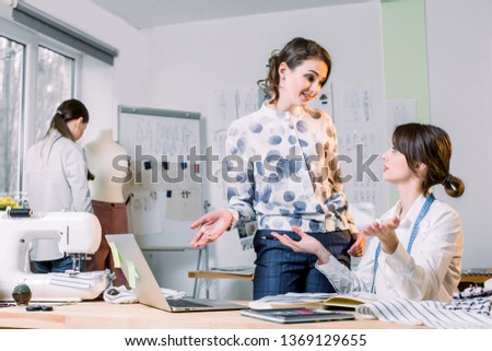 Young girls fashion designers using modern laptop and drawing sketches for new collection of clothes while working at table in studio. Woman dressmaker taking mannequin measurements with measure tape