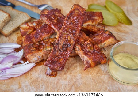 Close up Picture of american style bbq grilled pork spare ribs marinated in honey and served on wooden plate with slices of rustic bread, pickled cucumbers, chopped onions and french style hot mustard