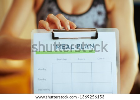Closeup on young woman in fitness clothes showing clipboard with meal plan in the modern living room.