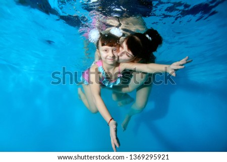 Happy mother kissing and hugging her young daughter under water in the pool. They play sports and smile on a blue background with their eyes open. Portrait. Horizontal view
