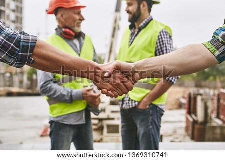 We did it! Close up photo of builders shaking hands against cheerful colleagues while working together at construction site. Team work.