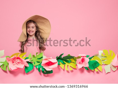 Portrait of a little girl in a summer hat on a pink background with paper flowers, place for text, summer advertising concept