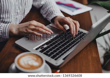 Female hands press laptop keys while sitting at wooden table with white cappuccino cup