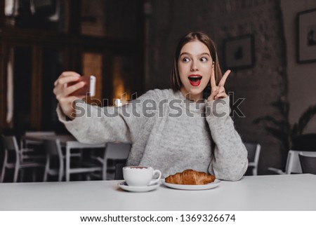 Energetic woman in gray sweater and red lipstick makes selfie. Portrait of girl showing peace sign in cafe with croissant on table