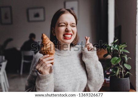 Picture of girl with red lips in cashmere outfit crossing fingers and wishing to eat croissant