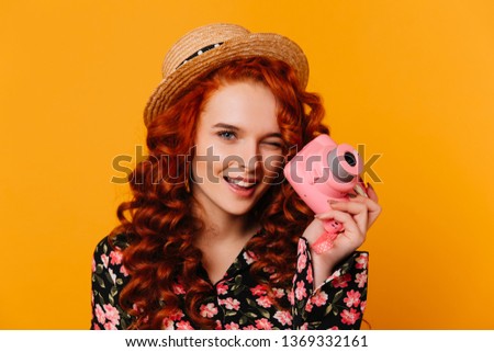 Red-haired girl in floral print blouse and blouse winks and holds pink camera on orange background