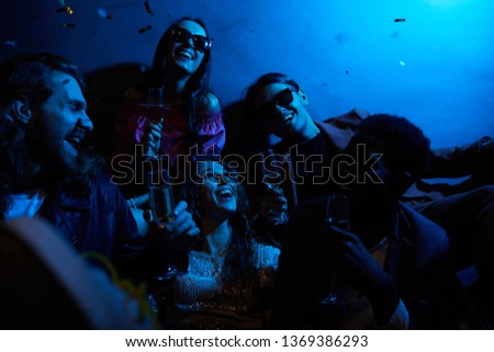 Group of positive young friends laughing together and drinking champagne in dark room with blue light, girl excited about falling confetti