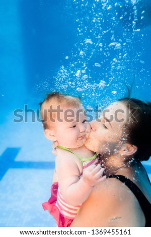 Mom kissing daughter are immersed in water, swimming under water in paddling pool. Diving baby. Learning infant child to swim. Young mother or swimming instructor and happy little girl.