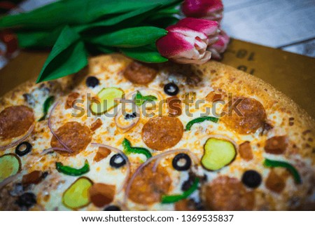 A whole pizza with olives, tomatoes, cheese, cucumbers on the table with red tulips. Pink tulips decorate pizza in a box.
