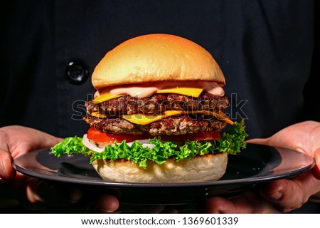 Male hands holding fresh delicious beef burger. Man in the black chef uniform serving homemade double patty burger with black angus beef, onion, tomato, cheese, lettuce, sauce on the black plate.