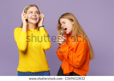 Two cheerful blonde twins sisters girls in vivid clothes listen music with headphones, sing song in microphone isolated on violet blue background. People family lifestyle concept. Mock up copy space