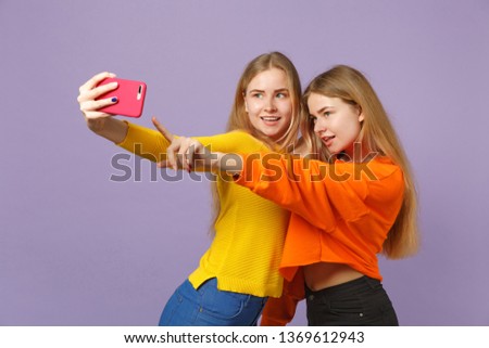 Two attractive young blonde twins sisters girls in vivid clothes doing selfie shot on mobile phone isolated on pastel violet blue wall background. People family lifestyle concept. Mock up copy space