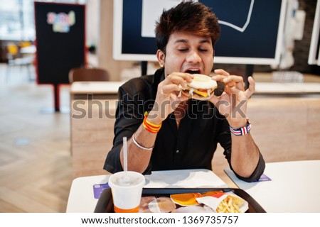 Stylish indian man sitting at fast food cafe and eating hamburger.