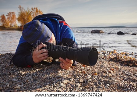A man photographs on a black long telephoto lens against the backdrop of the sea, lying on the rocks in an uncomfortable position.