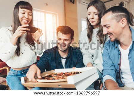 Good friends at home party. Group of four people eating pizza from delivery box and taking picture of food using mobile phone camera