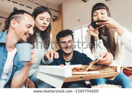 Friends at home party. Group of four people eating pizza from delivery box and taking photo of food using mobile phone camera