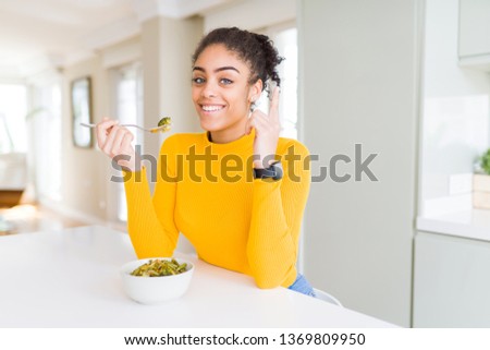 Young african american woman eating healthy green vegatables surprised with an idea or question pointing finger with happy face, number one Royalty-Free Stock Photo #1369809950