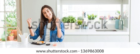 Wide angle picture of beautiful young woman eating delivery sushi very happy and excited doing winner gesture with arms raised, smiling and screaming for success. Celebration concept.