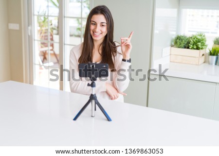 Beautiful young woman recording selfie video with smartphone webcam with a big smile on face, pointing with hand and finger to the side looking at the camera.