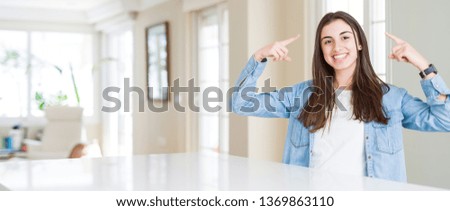 Wide angle picture of beautiful young woman sitting on white table at home Smiling pointing to head with both hands finger, great idea or thought, good memory