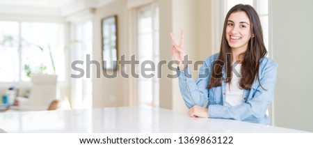 Wide angle picture of beautiful young woman sitting on white table at home smiling with happy face winking at the camera doing victory sign. Number two.