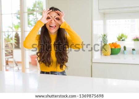 Young beautiful woman at home on white table doing ok gesture like binoculars sticking tongue out, eyes looking through fingers. Crazy expression.