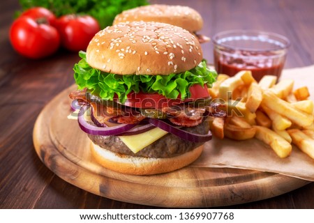 Hamburgers with fried bacon and sliced pickles  on a cutting board with french fries and ketchup