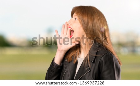 Young redhead woman shouting with mouth wide open to the lateral at outdoors