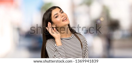 Teenager girl with striped shirt listening to something by putting hand on the ear at outdoors