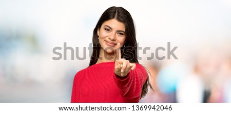 Teenager girl with red sweater showing and lifting a finger at outdoors