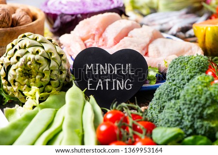 a black heart-shaped signboard with the text mindful eating, on a pile of different vegetables, such as French beans, cherry tomatoes, a head of broccoli, and some pieces of chicken in the background Royalty-Free Stock Photo #1369953164