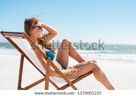 Smiling woman sunbathing on deck chair at beach. Attractive mature woman relaxing at seaside and looking the ocean. Young happy girl in bikini lying on a sun chair while wearing sunglasses. Royalty-Free Stock Photo #1370005235