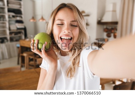 Image closeup of charming blond woman 20s wearing white t-shirt laughing and holding green apple while taking selfie photo in living room