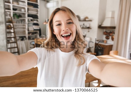Image closeup of content blond woman 20s wearing white t-shirt smiling while looking at camera and taking selfie photo in living room